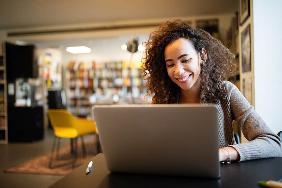 Client Center - Young Woman With Curly Hair and Tan Sweater Smiles as She Uses Her Laptop in a Colorful Coffee Shop